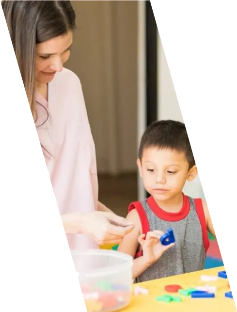 Una maestra acompaña a un niño mientras trabaja con piezas de colores sobre la mesa.
