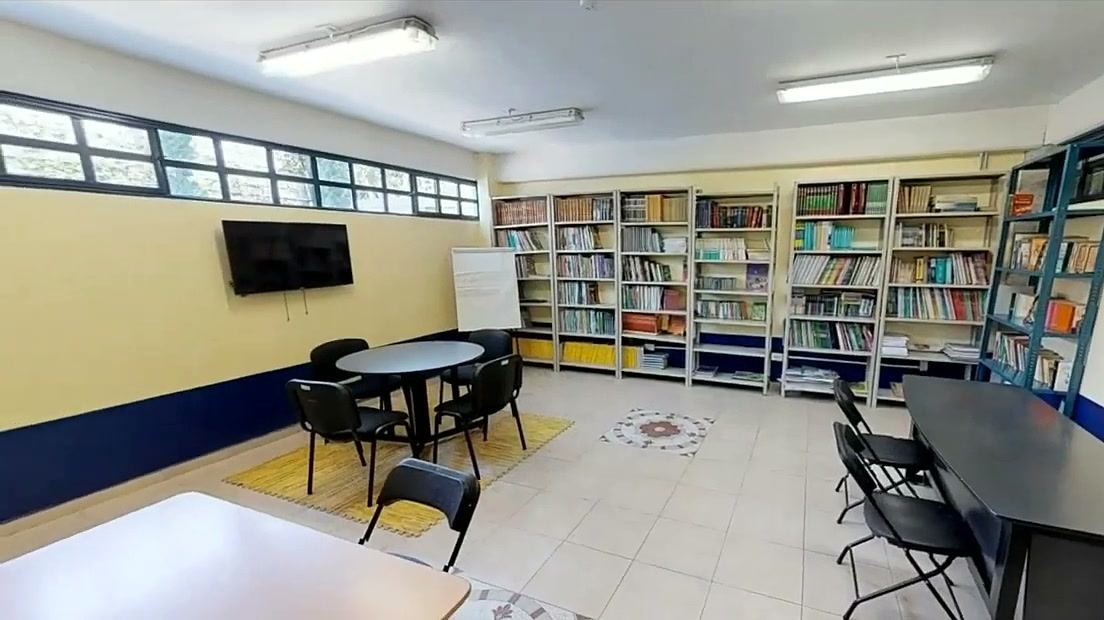 Salón con estanterías llenas de libros, una mesa redonda con sillas y una pantalla en la pared.