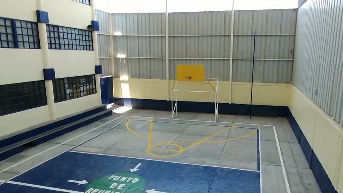 Patio interior con piso marcado como cancha y una canasta de básquetbol amarilla, rodeado de muros color crema y ventanas azules.