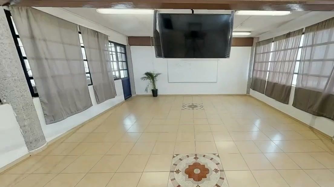 Salón amplio y vacío con piso de loseta, cortinas y una pantalla al fondo.
